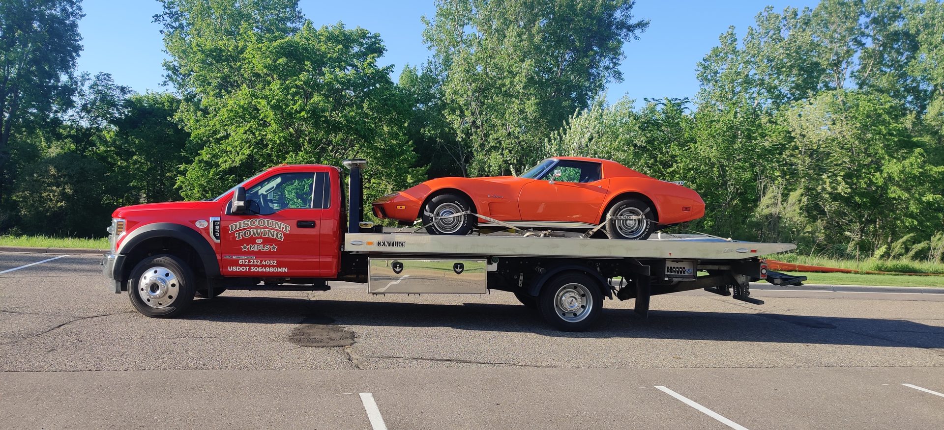 A red Discount Towing of MPLS flatbed truck loaded with a classic orange sports car in a sunlit parking lot
