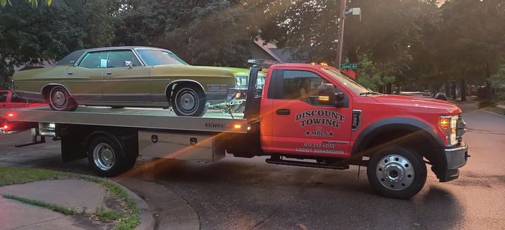 A red flatbed tow truck carrying a vintage green sedan on a city street in late-afternoon light