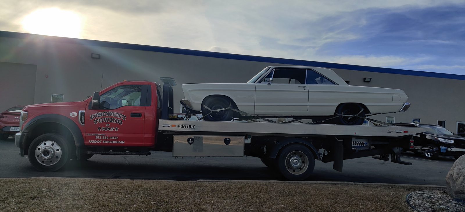 A red flatbed tow truck with a white classic coupe secured on its deck